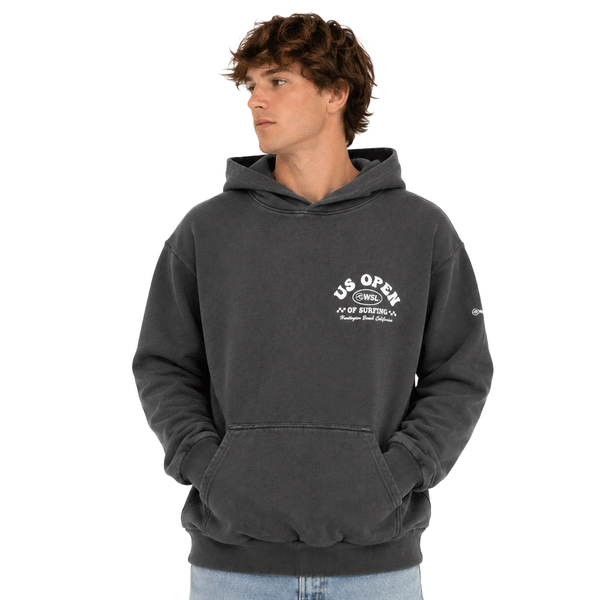 Front view of person wearing a charcoal heavyweight hooded sweatshirt featuring a white US Open of Surfing logo on the left chest. The pullover hoodie includes a kangaroo pocket, ribbed cuffs and waistband, and a vintage-washed finish, displayed on a clean white background.