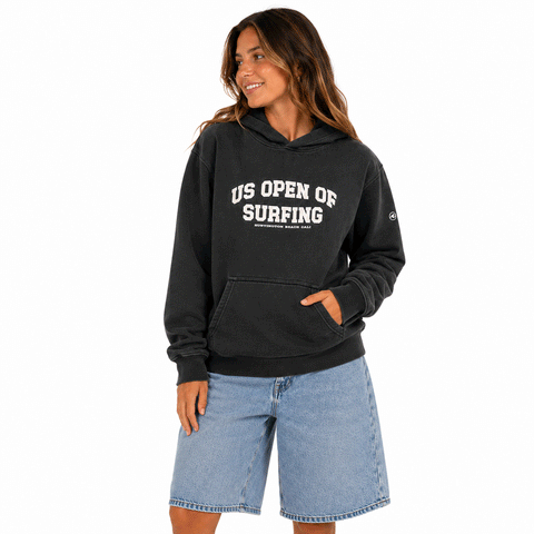 Woman wearing a washed black US Open of Surfing pullover hoodie with light denim shorts on a white background.