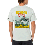 Back view of person wearing a light sage green men's T-shirt displayed on a clean white background. The shirt features a large, colorful "US Open of Surfing" graphic centered on the back, showcasing the Huntington Beach Pier, ocean waves, palm trees, and event branding in yellow, red, teal, and black. A small WSL logo is printed on the left sleeve. The design highlights the Huntington Beach, California surf event with a vibrant coastal illustration and retro-inspired typography.