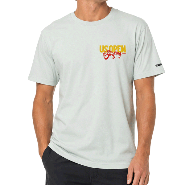front of person wearing a light sage green men's T-shirt displayed on a clean white background. The shirt features a small "US Open of Surfing" logo on the left chest in yellow and red. A subtle gray WSL logo is printed inside the collar, and a small black WSL logo appears on the right sleeve. The classic crewneck T-shirt has a relaxed fit and minimalist front design inspired by the Huntington Beach surf event.