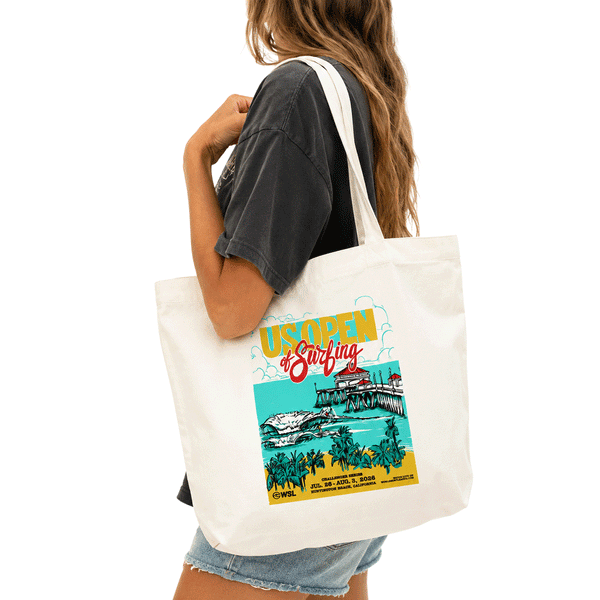 Woman carrying a cream canvas US Open of Surfing Huntington Beach tote bag with a colorful retro beach and pier graphic on a white background.